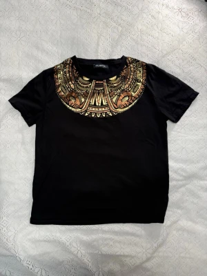 Svart t-shirt med metallicmönster Custo - Svart t-shirt från Custo Barcelona med korta ärmar och ett stort metallicmönster i guld och koppar över bröstet. Mönstret har coola detaljer med små nitar och ger en riktigt unik vibe. Tillverkad i mjuk bomull som känns skön mot huden.