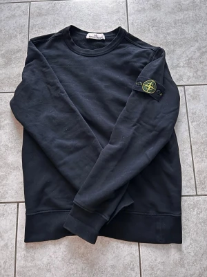Svart sweatshirt från Stone Island - Säljer en svart sweatshirt från Stone Island med den klassiska loggan på vänster ärm. Tröjan har rund halsringning, långa ärmar och ribbade muddar. Tillverkad i mjuk bomull för skön vardagsstil.