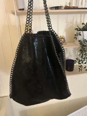 Svart axelväska med kedjedetaljer - Cool svart axelväska i glansig mockaimitation med grova kedjor längs kanterna och som axelband. Väskan har dragkedja upptill och flera innerfack, perfekt för att hålla ordning på dina grejer. Snygg och edgy design som sticker ut.