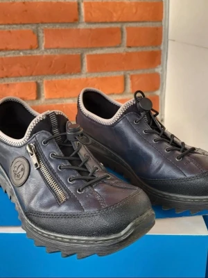 Rieker svarta sneakers i skinn med dragkedja 41 - Säljer ett par svarta sneakers från Rieker i skinn med coola detaljer som dragkedja på sidan, snörning med snabblås och grov sula. Skorna har rund tå och kontrastsöm runt öppningen. Perfekta för dig som vill ha en unik och bekväm stil.