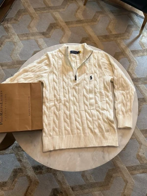 Vit kabelstickad tröja Polo Ralph Lauren - Säljer en vit kabelstickad tröja från Polo Ralph Lauren med halv dragkedja och broderad logga på bröstet. Tröjan har ribbade muddar och hög krage, perfekt för dig som gillar klassisk och clean stil.