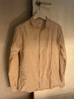Beige linneskjorta från RYVLS - Snygg beige skjorta från RYVLS Stockholm i mjukt linne. Klassisk krage, knäppning framtill och långa ärmar. Perfekt för dig som gillar en clean och enkel stil. Passar till allt och är superbekväm. Passar vanlig S och lite mindre S.