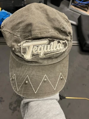 Grå keps med 'Tequila'-brodyr - Säljer en grå keps i bomull med broderad 'Tequila'-text framtill. Kepsen har vita sicksack-sömmar och små nitar på skärmen för extra edge. Justerbar rem baktill för perfekt passform. Perfekt accessoar för dig som vill sticka ut.