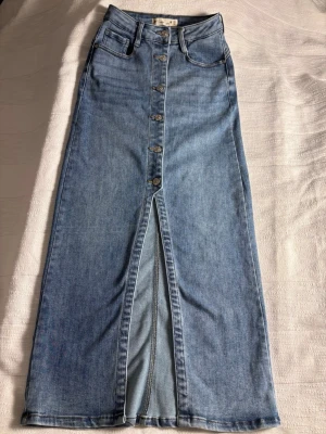 Lång jeanskjol från Denim Life XS - Säljer en snygg lång jeanskjol från Denim Life i ljusblå denim. Kjolen har knappar framtill, slits och klassiska fickor både fram och bak. Tillverkad i bomull och polyester med lite stretch för extra komfort.