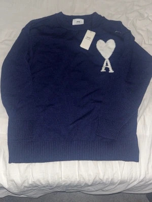 Mörkblå stickad tröja från Ami - Säljer en mörkblå stickad tröja från Ami Alexandre Mattiussi med vit hjärtdetalj och bokstaven A på bröstet. Klassisk rund halsringning och ribbade muddar vid ärmslut och nederkant. Perfekt för dig som gillar stilrena plagg med en twist.