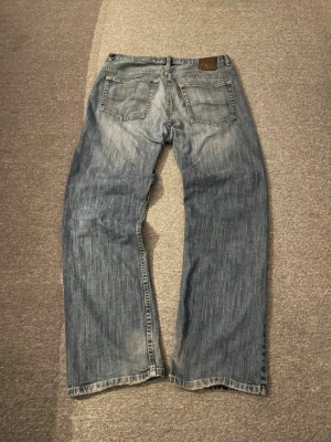 Lee Dungarees relaxed bootcut jeans - Säljer ett par blå Lee Dungarees relaxed bootcut jeans med klassisk femficksdesign och ljus tvätt. Jeansen har breda ben och avslappnad passform, perfekt för dig som gillar en chill och loose stil. Materialet är tåligt jeans med snygga slitningar och kontrastsömmar.