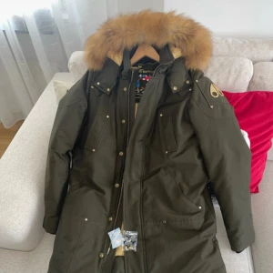 Olivgrön parkas Moose Knuckles päls - Säljer en olivgrön parkas från Moose Knuckles med stor fluffig päls runt huvan och guldfärgade detaljer. Jackan har flera fickor, dragkedja och knappar framtill samt ett broderat innerfoder. Perfekt för kalla dagar