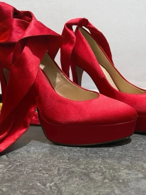 Röda klackar med platå och rosett - Snygga röda klackar 👠 med hög klack och platåsula. Skorna har en elegant rosett i siden som knyts runt ankeln för extra statement. Materialet är mjukt och ger en lyxig känsla, perfekta för att sticka ut.