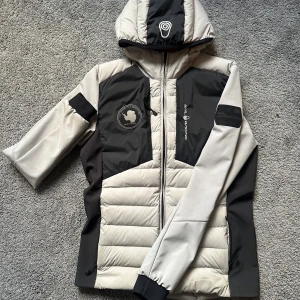 Sail Racing dunjacka med huva - Grym dunjacka från Sail Racing i svart, grått och vitt med röd insida. Jackan har huva, dragkedja och coola loggor på bröstet och ärmen. Fylld med 800 fill power dun från Allied Feather + Down. Perfekt för kalla dagar och snygg streetstil.