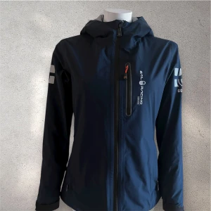 Mörkblå Sail Racing vindjacka - Snygg mörkblå vindjacka från Sail Racing med huva och vita tryck på bröstet och ärmen. Jackan har vattentätt GORE-TEX-material, hel dragkedja framtill och en bröstficka med zip. Perfekt för dig som gillar stilren och sportig look.#sailracing 46 cm bred och 61 cm lång