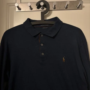 Mörkblå pikétröja från Ralph Lauren - Snygg mörkblå långärmad pikétröja från Ralph Lauren med klassisk krage, tre knappar och ikoniska broderade loggan på bröstet. Tröjan har en stilren look och är i ett mjukt bomullsmaterial som känns skönt mot huden.