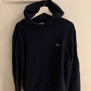 Lacoste Hoodie - Säljer en stilren mörkblå hoodie från Lacoste med klassisk krokodillogga på bröstet. Tröjan har huva, lång ärm och en stor magficka framtill. Perfekt för dig som gillar en enkel och snygg streetstil. Storlek XL men passar L