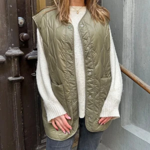 Olivgrön quiltad dunväst - Snygg olivgrön quiltad dunväst med tryckknappar och två stora fickor framtill. Västen har en rak, loose passform och är perfekt att slänga över en stickad tröja. Materialet är glansigt och lätt, vilket gör den både bekväm och trendig.