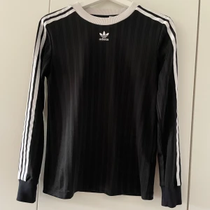 Svart långärmad Adidas tröja - Svart långärmad Adidas tröja med vita ränder längs ärmarna och broderad logga på bröstet. Tröjan har ribbad vit krage och är tillverkad i ett glansigt, sportigt material. Perfekt för en sportig och stilren look.