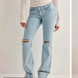 Ljusblå bootcut jeans med slitningar - Säljer ett par ljusblå jeans med bootcut-modell och snygga slitningar över knäna. Jeansen har klassisk femficksdesign, hög midja och är tillverkade i mjukt denimtyg. Perfekta för en avslappnad och trendig look.