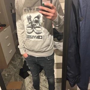 Grå hoodie från Converse med tryck - Säljer en grå hoodie från Converse med stort tryck framtill som föreställer ett par sneakers och Converse-loggan. Tröjan har huva, långa ärmar och ribbade muddar. Perfekt för dig som gillar streetstyle och vill ha något bekvämt och snyggt.