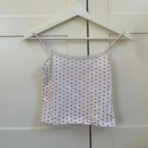 Vit linnetopp med röda hjärtan - Supersöt vit linnetopp med små röda hjärtan över hela tyget. Spaghettiband och spetskant upptill ger en gullig vibe. Perfekt för varma dagar eller som layering-piece. Tunt och mjukt bomullsmaterial.