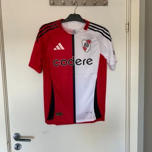 River Plate matchtröja Adidas M - River Plate fotbollströja från Adidas i slim fit. Tröjan är röd och vit med svart vertikal rand, klubbmärke på bröstet och sponsortryck. Klassiska Adidas-ränder på axlarna och tillverkad i lätt funktionsmaterial. Perfekt för dig som älskar argentinsk fotboll.
