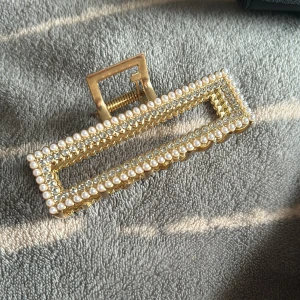 Guldig hårklämma med pärlor och strass - Rektangulär hårklämma i guldmetall dekorerad med små vita pärlor och glittrande strass. Klämman har en öppen design med raka linjer och ger en lyxig känsla till din frisyr. Perfekt accessoar för att lyfta din look med extra bling.