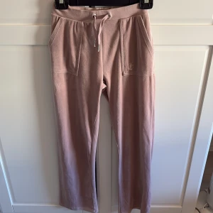 Rosa mjukisbyxor från Juicy Couture - Mysiga rosa mjukisbyxor från Juicy Couture med raka ben, snörning i midjan och stora fickor både fram och bak. Byxorna har en mjuk och skön känsla, perfekt för chill dagar. Klassisk Juicy Couture-logga broderad på ena fickan framtill.