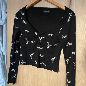 Svart blommig croppad topp Even&Odd - Svart croppad topp från Even&Odd med vita blommiga detaljer. Toppen har lång ärm, v-ringning och ribbad struktur. Snygg vågig kant nedtill och lätt rynkning vid bröstet ger en trendig look. Perfekt för dig som gillar stilrena och coola plagg. Nästan aldrig använd pga. Att den är för stor för mig.