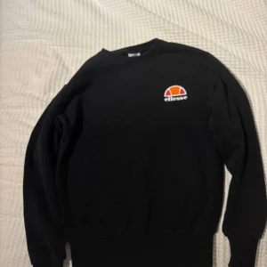 Svart sweatshirt från Ellesse - Svart sweatshirt från Ellesse med klassisk logga i vitt, orange och rött på bröstet. Tröjan har rund halsringning och långa ärmar. Perfekt för en chill och sportig stil.Ellesse sweatshirt/långärmad säljs då jag inte längre använder den skicket är mycket bra inga skavanker. Storlek XS 9/10 skick