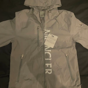 Grå vindjacka från Moncler med huva - Snygg grå vindjacka från Moncler med huva och dragkedja framtill. Jackan har tre fickor med dragkedja, stor Moncler-logga på bröstet och klassisk patch på ärmen. Perfekt för dig som vill ha en stilren och sportig look.