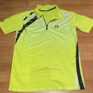 FZ Forza - Gul pikétröja / Badminton  - Sportig gul pikétröja från FZ Forza med svart och vit detalj på axeln och sidan. Tröjan har kort ärm, krage och half zip framtill. Tillverkad i lätt och snabbtorkande polyester, perfekt för träning eller tennis.