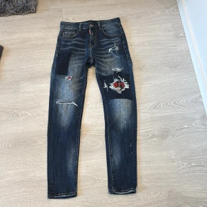 Dsquared2 slim jeans med slitningar - Mörkblå slim jeans från Dsquared2 med coola slitningar, röda patch-detaljer och färgstänk. Klassisk femficksmodell med låg midja och smal passform. Jeansmaterialet är robust och har en snygg tvättad look. Perfekta för dig som gillar streetstyle och unika detaljer.