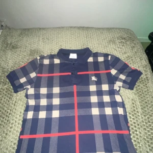 Rutig kortärmad Burberry skjorta - Säljer en snygg kortärmad skjorta från Burberry med klassiskt rutigt mönster i marinblått, beige och röda linjer. Skjortan har krage, knappar framtill och är tillverkad i mjuk bomull. Perfekt för dig som vill sticka ut med en ikonisk stil.