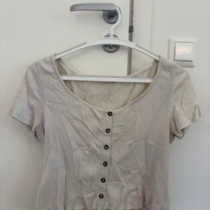 Beige/vit blus med knappar - Snygg beige blus med dekorativa knappar framtill. Modellen har korta ärmar och en rundad halsringning. Perfekt för en avslappnad och trendig look. TVÄTTAS OCH STRYKS INNAN DEN SKICKAS