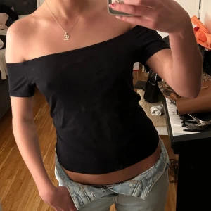 Svart offshoulder topp i bomull - Säljer en svart offshoulder topp med korta ärmar. Sitter tight och har en stilren, enkel design utan mönster eller detaljer. Perfekt att styla med jeans eller kjol för en clean look. Gjord i mjukt bomullsmaterial som känns skönt mot huden.