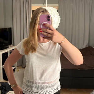 Vit blus med spets från Vero Moda - Supersöt vit blus från Vero Moda i bomull med korta ärmar och rund hals. Nedtill finns en bred spetskant och virkade detaljer som ger en romantisk vibe. Perfekt till jeans eller kjol för pigga upp outfiten lite extra!