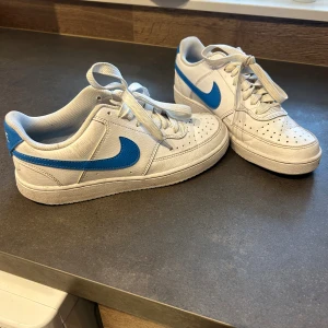 Nike Air Force 1 vita/blå sneakers - Säljer ett par klassiska Nike Air Force 1 sneakers i vitt skinn med blå swoosh. Lågt skaft, perforerad tå och snörning framtill. Perfekta för dig som gillar en clean och sportig look med en färgklick. Endast använda 2ggr så nyskick!