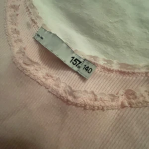 Ljusrosa ribbad linnetopp med spets - Söt ljusrosa linnetopp med ribbad struktur och spetskant runt halsringning och ärmhål. Perfekt för en enkel och gullig look. Toppen är ärmlös och har en klassisk passform.