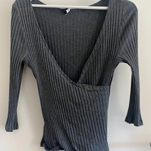 Grå ribbad omlottopp från 77FLEA - Säljer en mörkgrå omlottopp från 77FLEA i storlek L. Toppen har ribbad struktur, djup v-ringning och trekvartsärmar. Materialet är mjukt och stretchigt. Nedre kanten har en lätt volang för extra detalj.