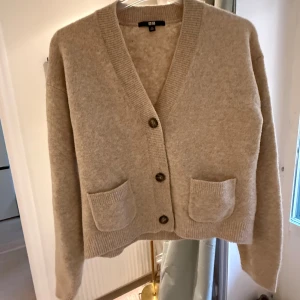 Beige kofta från Uniqlo med fickor - Mysig beige kofta från Uniqlo med v-ringning, tre knappar framtill och två praktiska fickor. Koftan är stickad och har ribbade detaljer vid ärmslut, fickor och nederkant. Perfekt för lager-på-lager och chill stil.