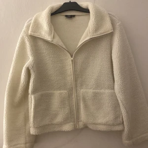 Vit teddyjacka från lager 157, storlek M - Mysig vit teddyjacka från lager 157 i storlek M. Jackan har dragkedja framtill, två stora fickor och en klassisk krage. Perfekt för kyliga dagar när du vill vara både varm och snygg. Materialet är fluffigt
