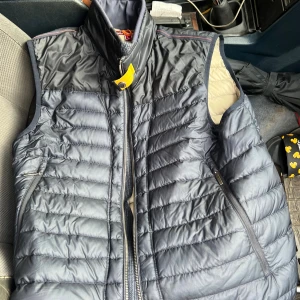 Mörkblå dunväst från Parajumpers L - Snygg mörkblå dunväst från Parajumpers Day Tripper med quiltad design, hög krage och dragkedja framtill. Västen har två sidofickor med dragkedja och en ikonisk gul detalj vid halsen. Insidan är beige och materialet är lätt och smidigt. KAN ÄVEN BYTAS MOT PARAJUMPER I ANNAN FÄRG!