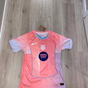 Barça x Nike rosa fotbollströja - Snygg FC Barcelona fotbollströja från Nike i rosa och ljusblå toner. Tröjan har korta ärmar, klubbmärke och sponsorlogga framtill samt tunna blå detaljer längs kanterna. Tillverkad i lätt och ventilerande material, perfekt för match eller träning.