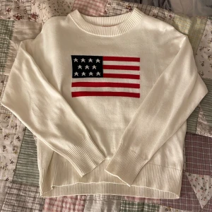 Vit stickad tröja med USA-flagga RYVLS - Vit stickad tröja från RYVLS med amerikansk flagga på bröstet. Tröjan har ribbade muddar vid ärmslut och nederkant samt rund halsringning. Materialet är en mix av bomull och akryl, vilket gör den både mjuk och bekväm. Perfekt för dig som gillar klassisk och stilren design.