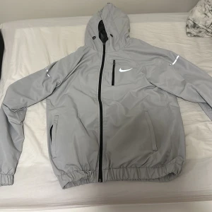 Grå Nike vindjacka med huva - Grå vindjacka från Nike med huva och svarta dragkedjor. Jackan har reflekterande detaljer på ärmarna och bröstet, meshfoder på insidan och två sidofickor. Perfekt för dig som gillar sportig stil och vill ha något lätt och snyggt.