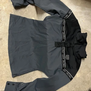 Svart långärmad tröja Maya Delorez XS - Svart långärmad tröja från Maya Delorez i storlek XS. Tröjan har hög krage, knappar framtill och detaljer med logga längs axlar och ärmslut. Snygg och sportig design som passar perfekt till ridning eller andra aktiviteter.