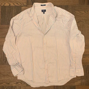 Randig skjorta från GANT i bomull - Riktigt snygg skjorta från GANT i storlek L med ljusblå, vita och röda ränder. Klassisk krage, broderad logga på bröstet och knappar framtill. Tillverkad i mjuk bomull och har lång ärm med manschett. Perfekt för dig som gillar stilrena och fräscha plagg.  Vid funderingar fråga på! 