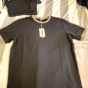 Svart t-shirt med beige detaljer från GRAZ - Snygg svart t-shirt från GRAZ med beige ribbad krage och ärmslut. Enkel och stilren design som passar perfekt till jeans eller shorts. Tillverkad i mjuk bomull för en skön känsla hela dagen.