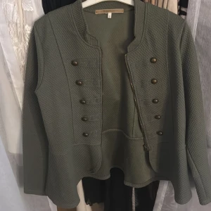 Olivgrön kavaj med dekorativa knappar - Snygg olivgrön kavaj med strukturerat tyg och dekorativa bronsfärgade knappar framtill. Jackan har lång ärm, markerad midja och dragkedja i sidan. Perfekt för dig som vill ha en unik och trendig look.