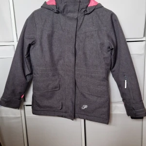 Grå parkajacka från On The Peak - Säljer en grå parkajacka från On The Peak med huva och rosa detaljer på insidan. Jackan har flera praktiska fickor, dragkedja och kardborre vid ärmslut. Perfekt för kyliga dagar och snygg till vardagslooken.