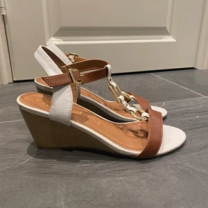 Bruna och vita sandaler med kilklack - Snygga sandaler från Marco Tozzi i brunt och vitt skinn med kilklack i trälook. Skorna har öppen tå, remmar med guldfärgade metalldetaljer och spänne vid ankeln. Perfekta för sommarens alla tillfällen och ger en fräsch och stilren look.