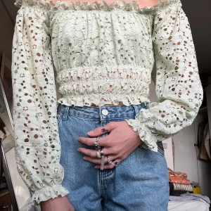 Ljusgrön offshoulder blus med spets - Superfin ljusgrön offshoulder blus med broderad spets och hålmönster. Croppad modell med smockad midja och volangdetaljer längs kanterna. Långa puffärmar med resår vid ärmslut. Perfekt för vår och sommar!