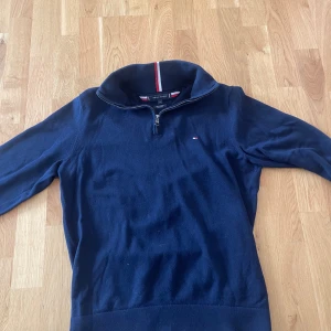 Marinblå half zip-tröja Tommy Hilfiger - Klassisk marinblå tröja från Tommy Hilfiger med half zip och hög krage. Liten broderad logga på bröstet och ribbade muddar. Storlek M